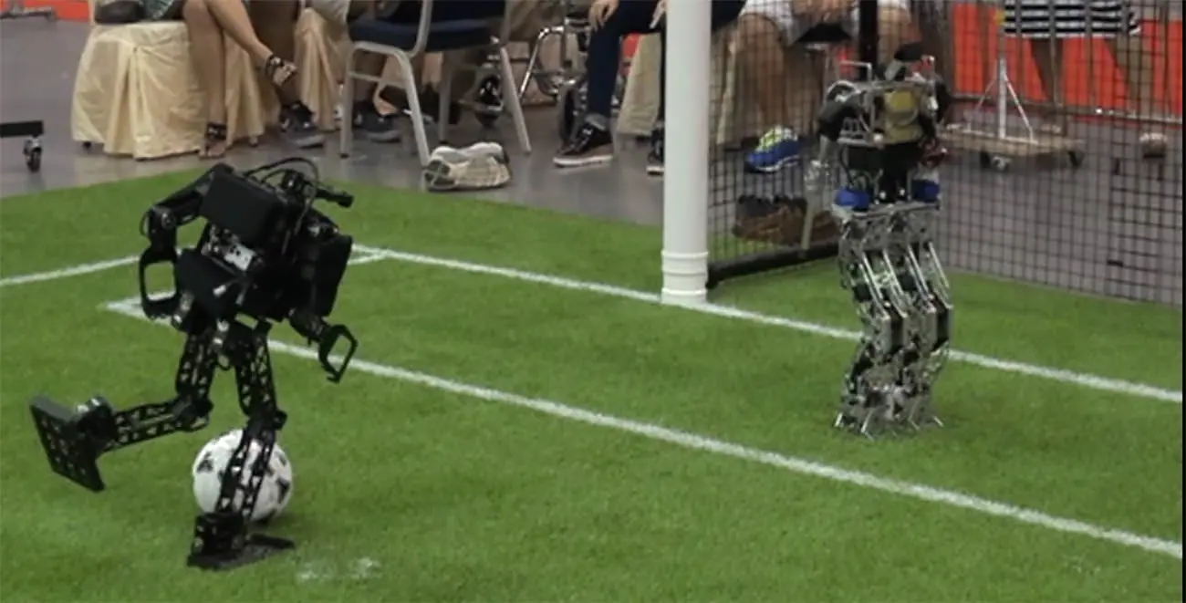Two humanoid robots on a RoboCup soccer field, one mid-kick next to the ball in front of a goal.